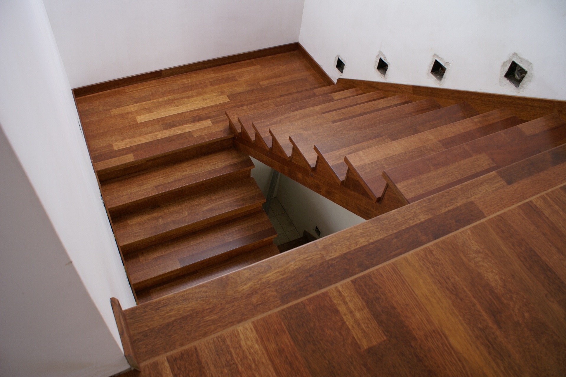 Widok z góry na drewniane schody z ciemnego drewna, z widocznymi fragmentami podłogi i ściany, gdzie widać przygotowane otwory na punkty świetlne.