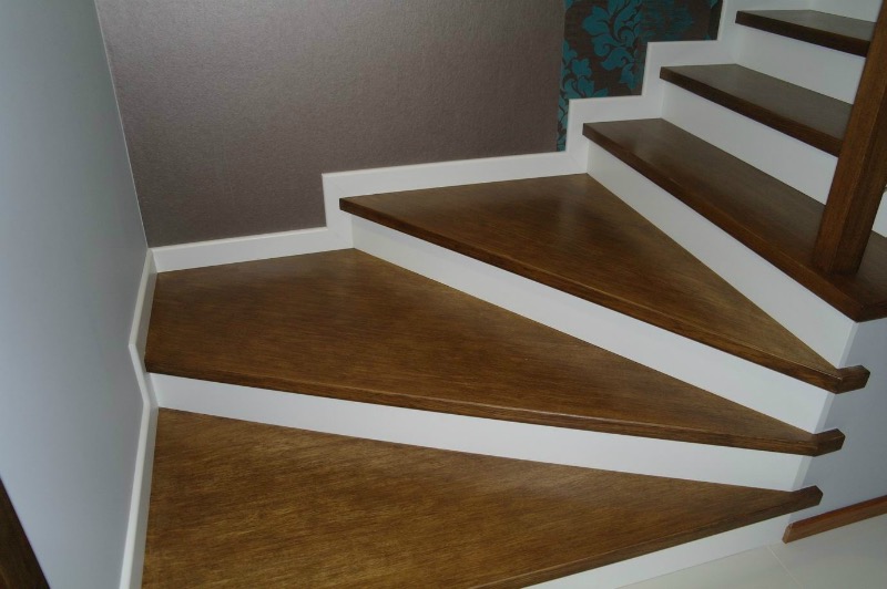 Drewniane schody z białymi podstopnicami, widoczne z dołu, z ciemnobrązowymi stopniami i wzorzystą tapetą w tle.