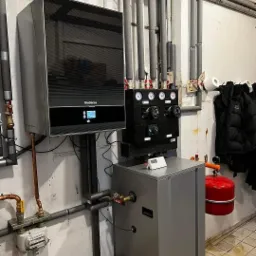 Nowoczesna instalacja grzewcza z kotłem gazowym Buderus, czarnym modułem sterującym, szarym zasobnikiem ciepłej wody Apriso i licznymi rurami zabezpieczonymi izolacją termiczną, zamontowana...