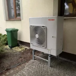 Biała pompa ciepła Toshiba zamontowana na metalowych wspornikach przy ścianie budynku w Łodzi, obok stoi zielony kosz na śmieci, a w tle widoczne okno.