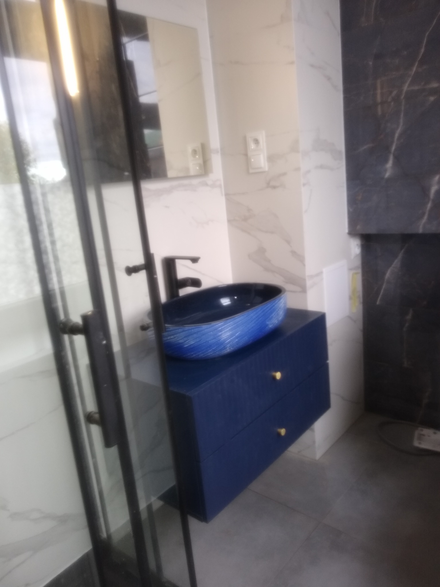 Niebieska, owalna umywalka nablatowa z efektem fal na szafce z dwiema szufladami i złotymi uchwytami, na tle marmurowych płytek w łazience z czarną armaturą i kabiną prysznicową ze szklanymi drzwiami.