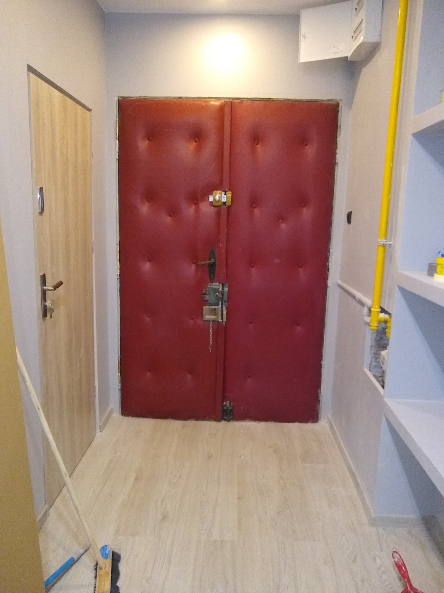 Korytarz po remoncie z nową podłogą z paneli w odcieniach beżu, czerwonymi drzwiami wejściowymi z tapicerowanym wykończeniem i jasnoszarymi ścianami; widoczna rura gazowa w kolorze żółtym.