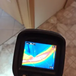 Kamera termowizyjna pokazuje rozkład temperatur w rogu pomieszczenia, z widocznym mostkiem termicznym przy podłodze i ścianie, na tle beżowych płytek podłogowych i fragmentu drzwi.