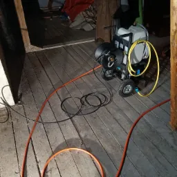 Przenośne urządzenie do udrażniania rur z bębnem na wąż, zbiornikiem na wodę i wężem w kolorze pomarańczowym, leżącym na drewnianej podłodze poddasza.