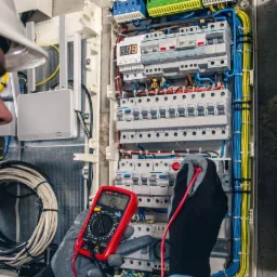 Elektryk w kasku, mierzący napięcie w skrzynce rozdzielczej za pomocą multimetru z czerwonymi przewodami, widoczne bezpieczniki i okablowanie.