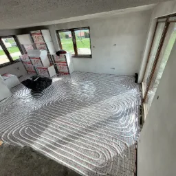 Białe rury ogrzewania podłogowego ułożone w pętle na folii z czerwoną siatką w surowym wnętrzu z oknami, widoczne kartony izolacyjne i czarny worek.