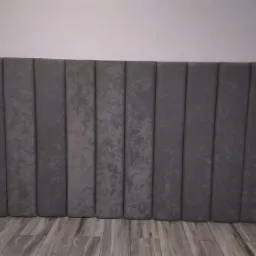 Szary, tapicerowany zagłówek łóżka z pionowymi przeszyciami, ustawiony na podłodze z paneli imitujących drewno.