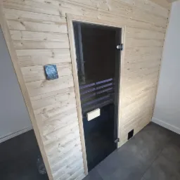 Wnętrze sauny z drewnianym wykończeniem, szklanymi drzwiami i małą płytką dekoracyjną na ścianie obok wejścia.