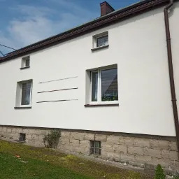 Ocieplony dom jednorodzinny z białą elewacją, widoczne ślady po taśmie malarskiej, brązowe rynny i podmurówka z kamienia, częściowo widoczne rusztowanie i samochód dostawczy w tle.