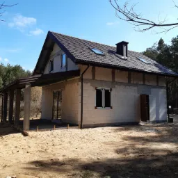 Dom w trakcie budowy z ciemnobrązowym dachem z dachówki, widoczne okna dachowe i komin, surowy stan elewacji z betonu komórkowego, zadaszenie tarasu z betonowymi słupami, piaszczyste podłoże.