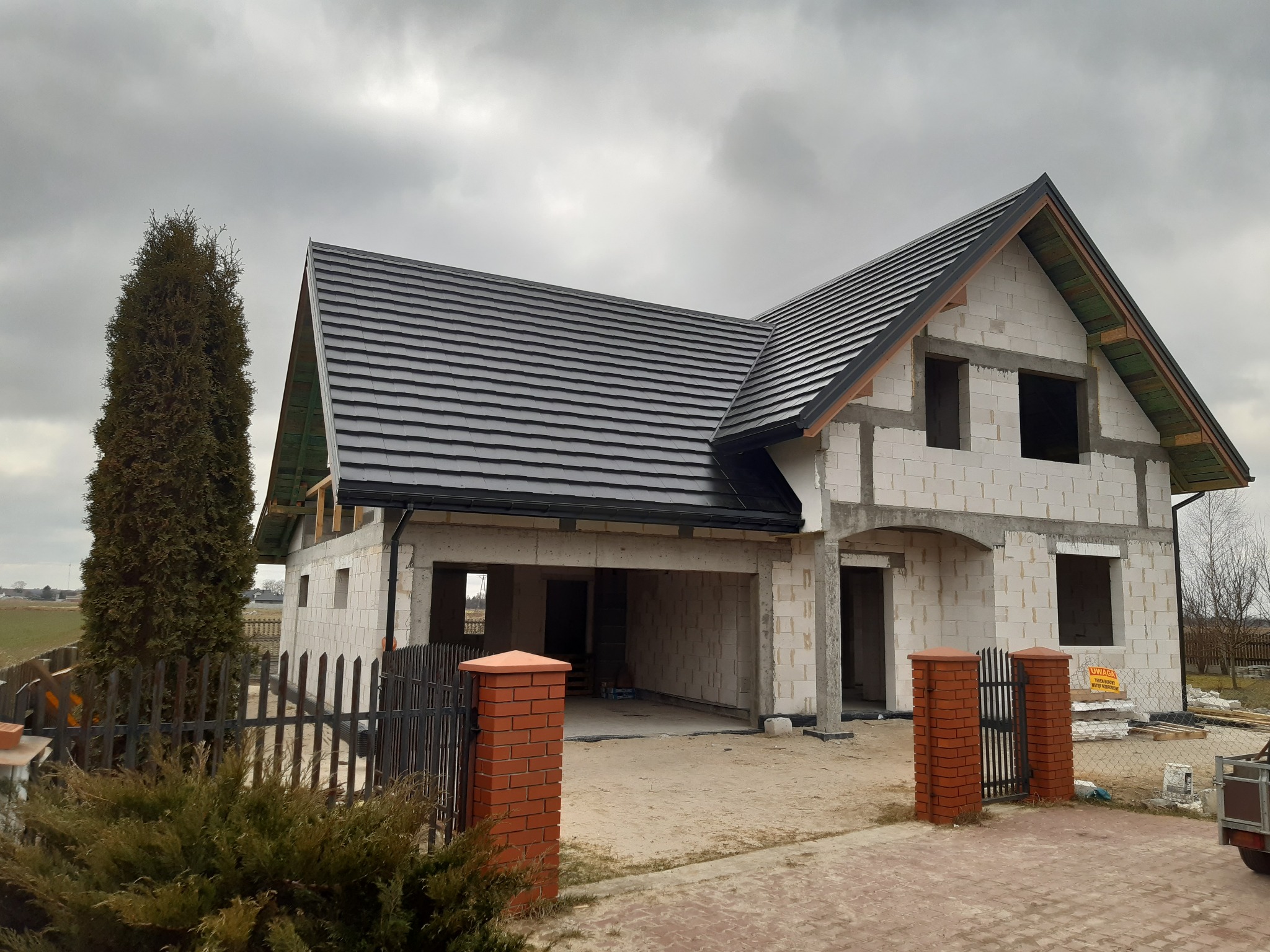 Surowy stan domu jednorodzinnego z nowym, czarnym dachem z dachówki ceramicznej, widok z zewnątrz, w tle pochmurne niebo.