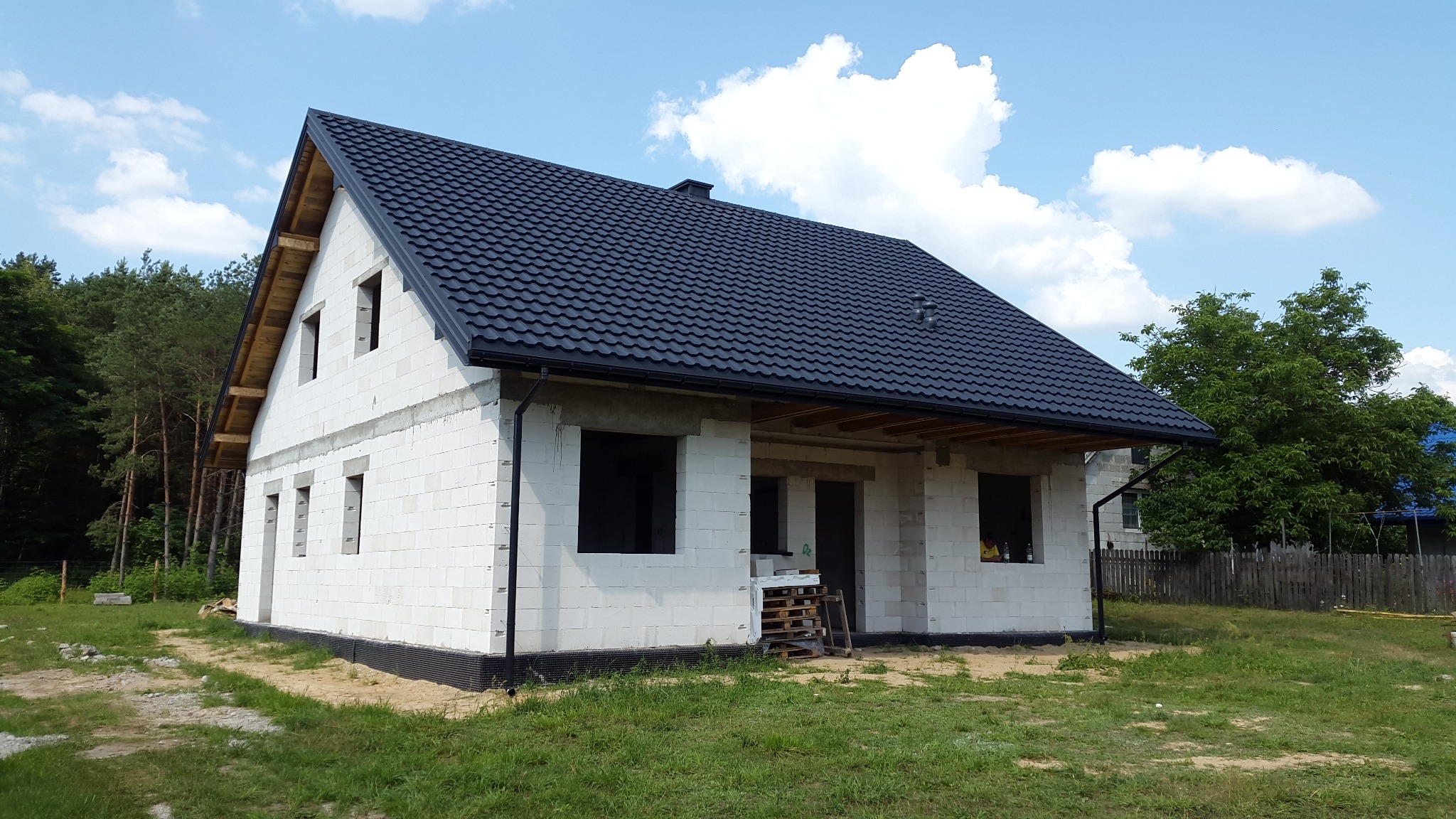 Dom w stanie surowym zamkniętym z czarnym dachem z blachodachówki, widoczne rynny, ściany z białego bloczka, okna i drzwi bez stolarki, drewniana więźba dachowa, w tle drzewa i błękitne niebo.