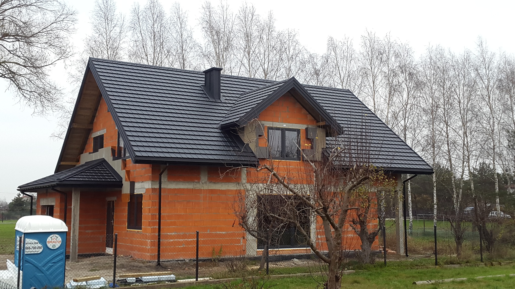 Nowy, grafitowy dach z dachówki ceramicznej na surowym budynku z czerwonej cegły, widoczna rynna i komin, w tle drzewa bez liści i toaleta przenośna.