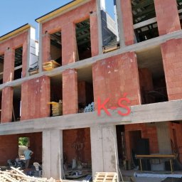 Krzysztof Śliwa Docieplenia i Remonty - Wznoszenie murów z czerwonej cegły na nowym trzypiętrowym budynku z widocznymi elementami konstrukcyjnymi i stertami materiałów budowlanych wewnątrz.