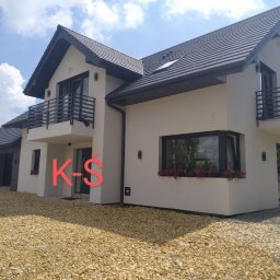 Krzysztof Śliwa Docieplenia i Remonty - Elewacja dwukondygnacyjnego domu z balkonami, wykończona jasnym tynkiem, ciemna dachówka i stolarka okienna, widok z poziomu gruntu na podjazd z kamieni.