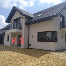 Elewacja dwukondygnacyjnego domu z balkonami, wykończona jasnym tynkiem, ciemna dachówka i stolarka okienna, widok z poziomu gruntu na podjazd z kamieni.