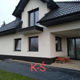 Krzysztof Śliwa Docieplenia i Remonty - Boczny widok na dom z wykończoną elewacją w kolorze beżowym z czarnymi akcentami, widoczne niedociągnięcia na ścianie i materiały budowlane przed wejściem.