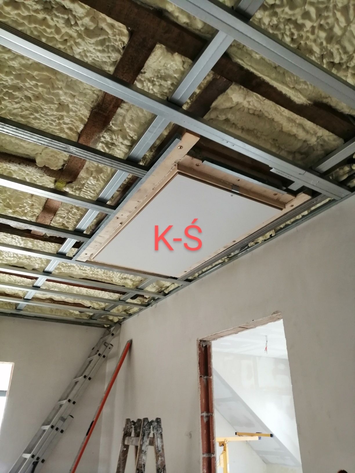 Sufit z ociepleniem pianką PUR, widoczne metalowe profile, drewniana rama otworu rewizyjnego, częściowo wykończone pomieszczenie z drabinami.