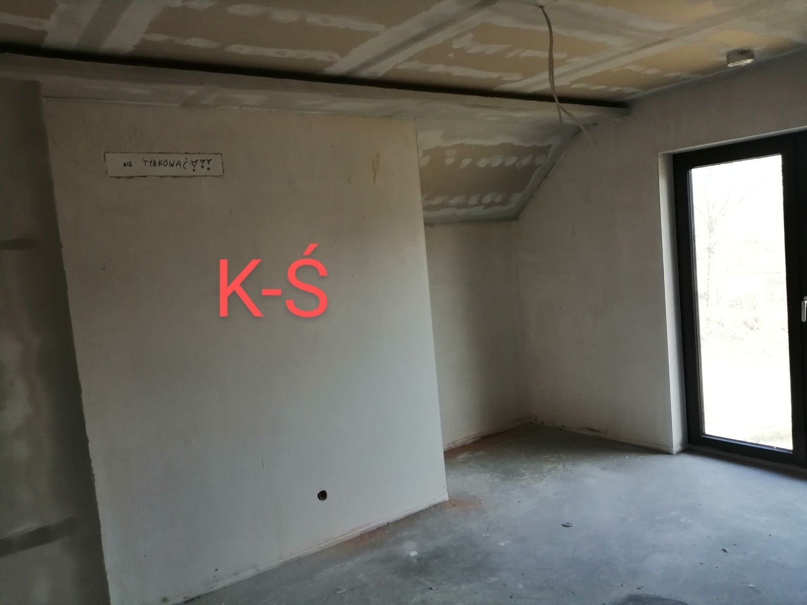 Surowy stan wnętrza pokoju na poddaszu z oknem, ściany przygotowane do malowania, sufit z płyt kartonowo-gipsowych przed szpachlowaniem, na ścianie napisane 'Nie tynkować'.