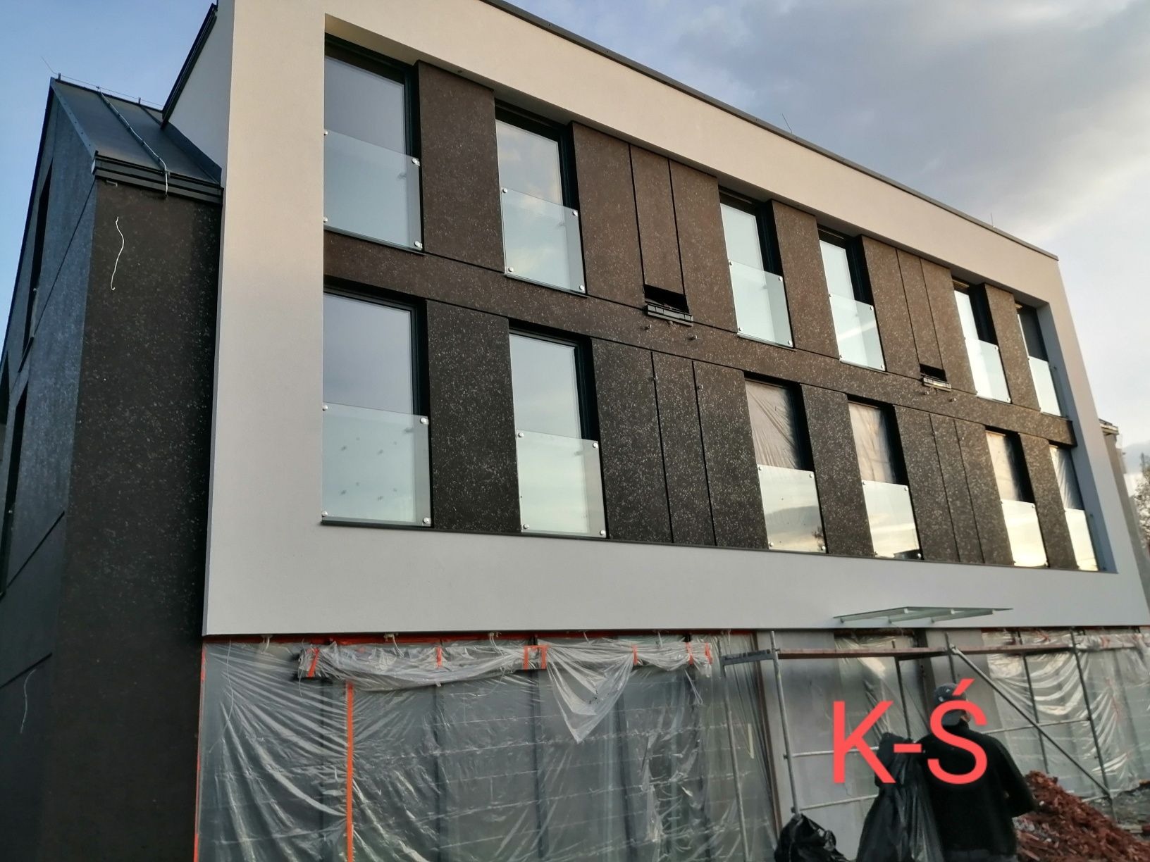 Nowoczesny budynek z czarną elewacją w trakcie ocieplania, zabezpieczony folią ochronną, widoczny fragment rusztowania i pracownik w ciemnym ubraniu.