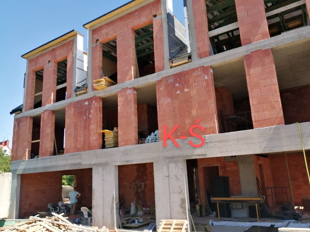 Wznoszenie murów z czerwonej cegły na nowym trzypiętrowym budynku z widocznymi elementami konstrukcyjnymi i stertami materiałów budowlanych wewnątrz.