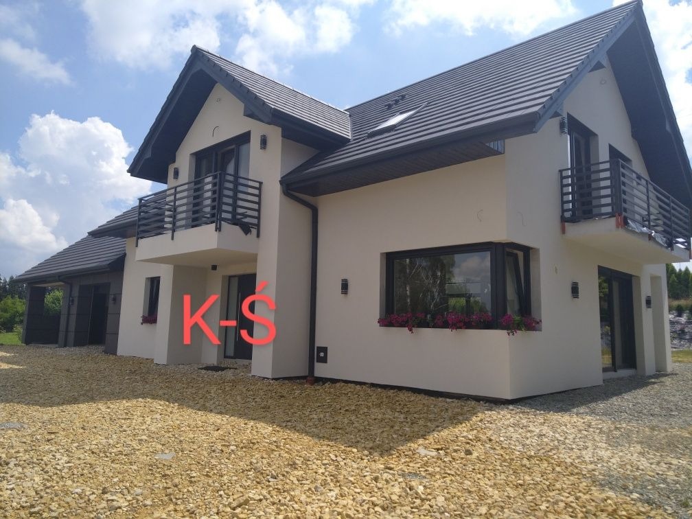 Elewacja dwukondygnacyjnego domu z balkonami, wykończona jasnym tynkiem, ciemna dachówka i stolarka okienna, widok z poziomu gruntu na podjazd z kamieni.