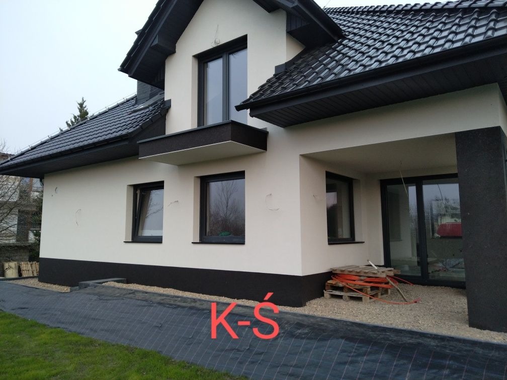 Boczny widok na dom z wykończoną elewacją w kolorze beżowym z czarnymi akcentami, widoczne niedociągnięcia na ścianie i materiały budowlane przed wejściem.