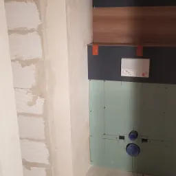 Ściana łazienki w trakcie remontu: widoczne bloczki, fragment płyty gipsowej z zaznaczonymi otworami na instalację, fragment zabudowy z płyt w kolorze drewna i grafitu oraz schowana spłuczka.