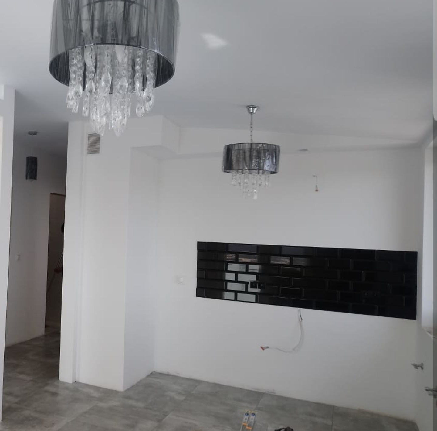 Wnętrze w trakcie prac wykończeniowych, z dwoma nowoczesnymi żyrandolami z kryształkami, ułożona czarna, błyszcząca mozaika na ścianie i widoczne kable, podłoga wyłożona szarymi płytkami...