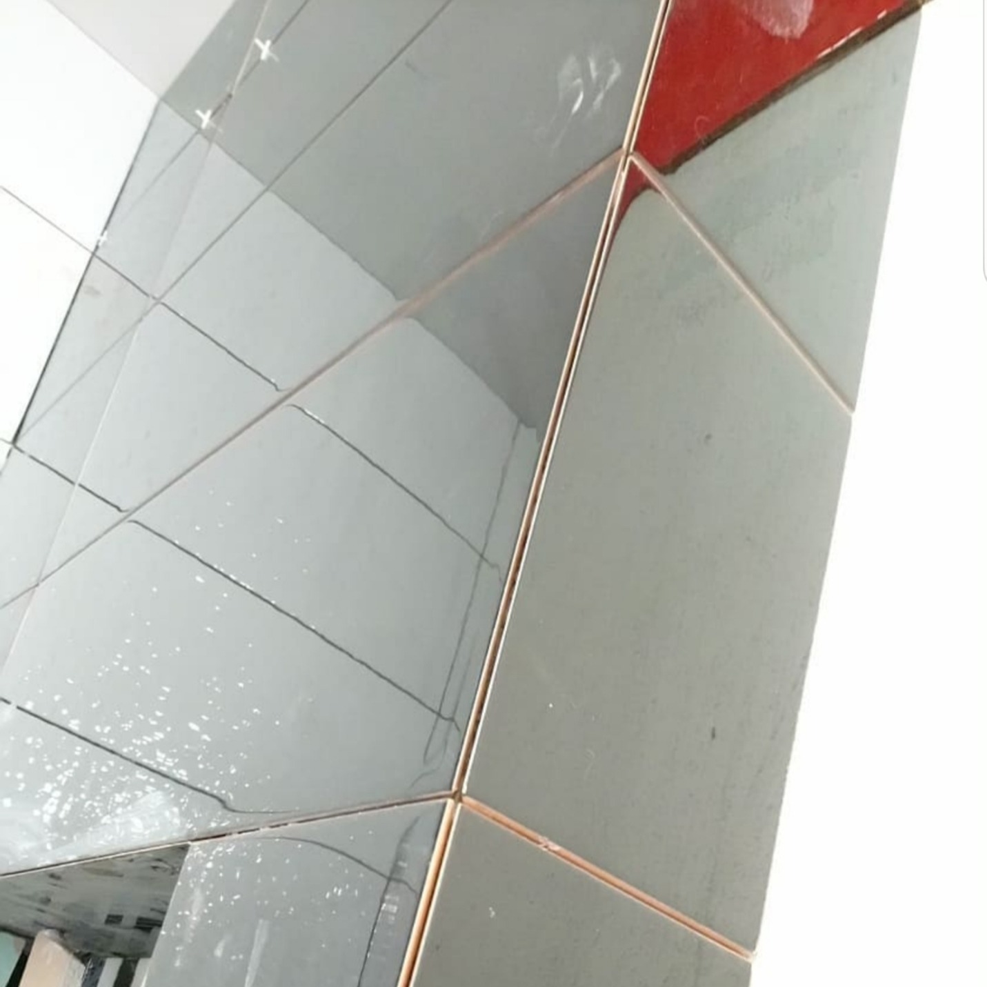 Świeżo ułożone, błyszczące, szare płytki na ścianie, z widocznymi krzyżykami dystansowymi i smugami po kleju, pod kątem, z fragmentem czerwonej ściany w górnym rogu.