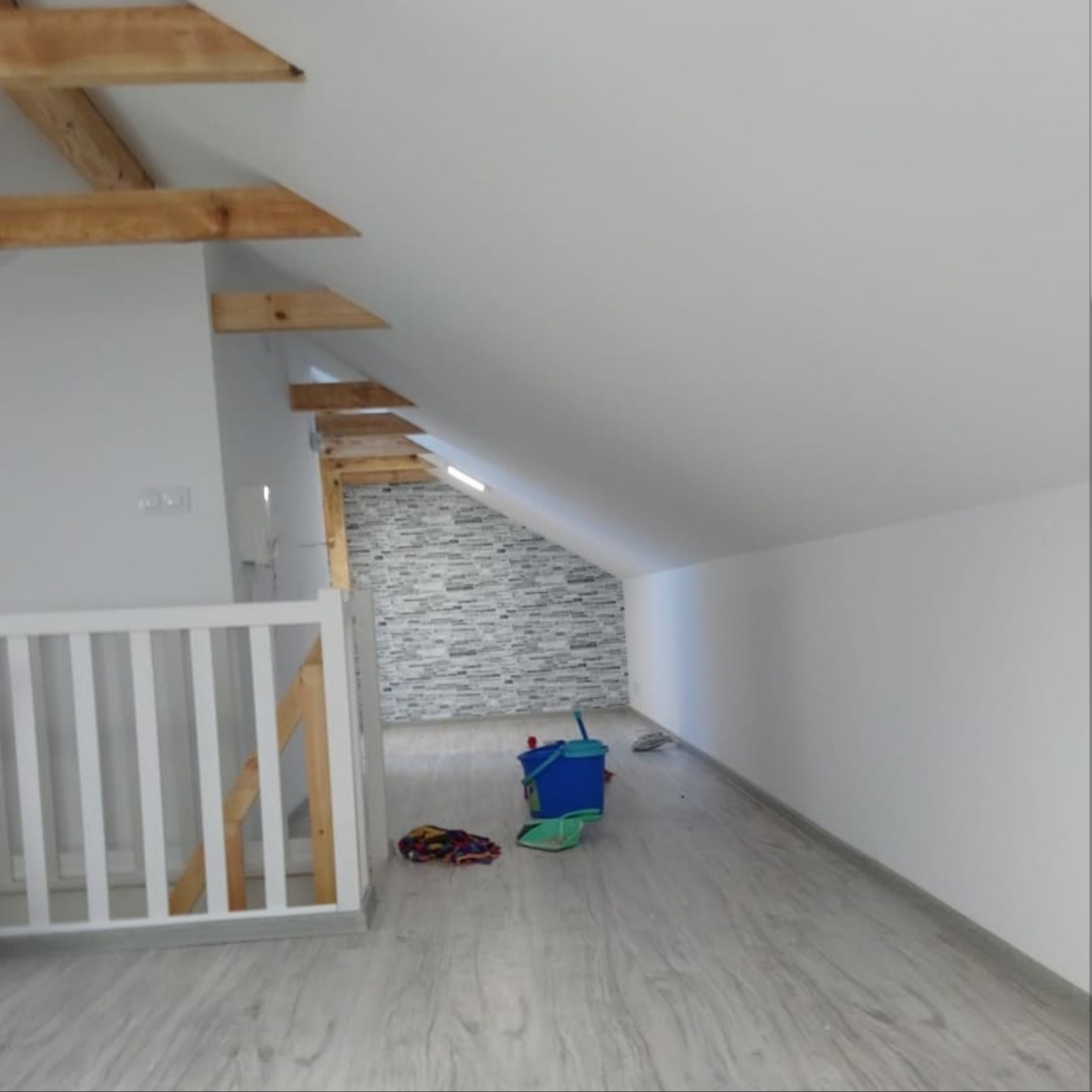 Poddasze z drewnianą konstrukcją dachu, pomalowanymi na biało ścianami i szarą tapetą imitującą kamień na jednej ze ścian, widoczne narzędzia i akcesoria malarskie, podłoga wyłożona panelami.