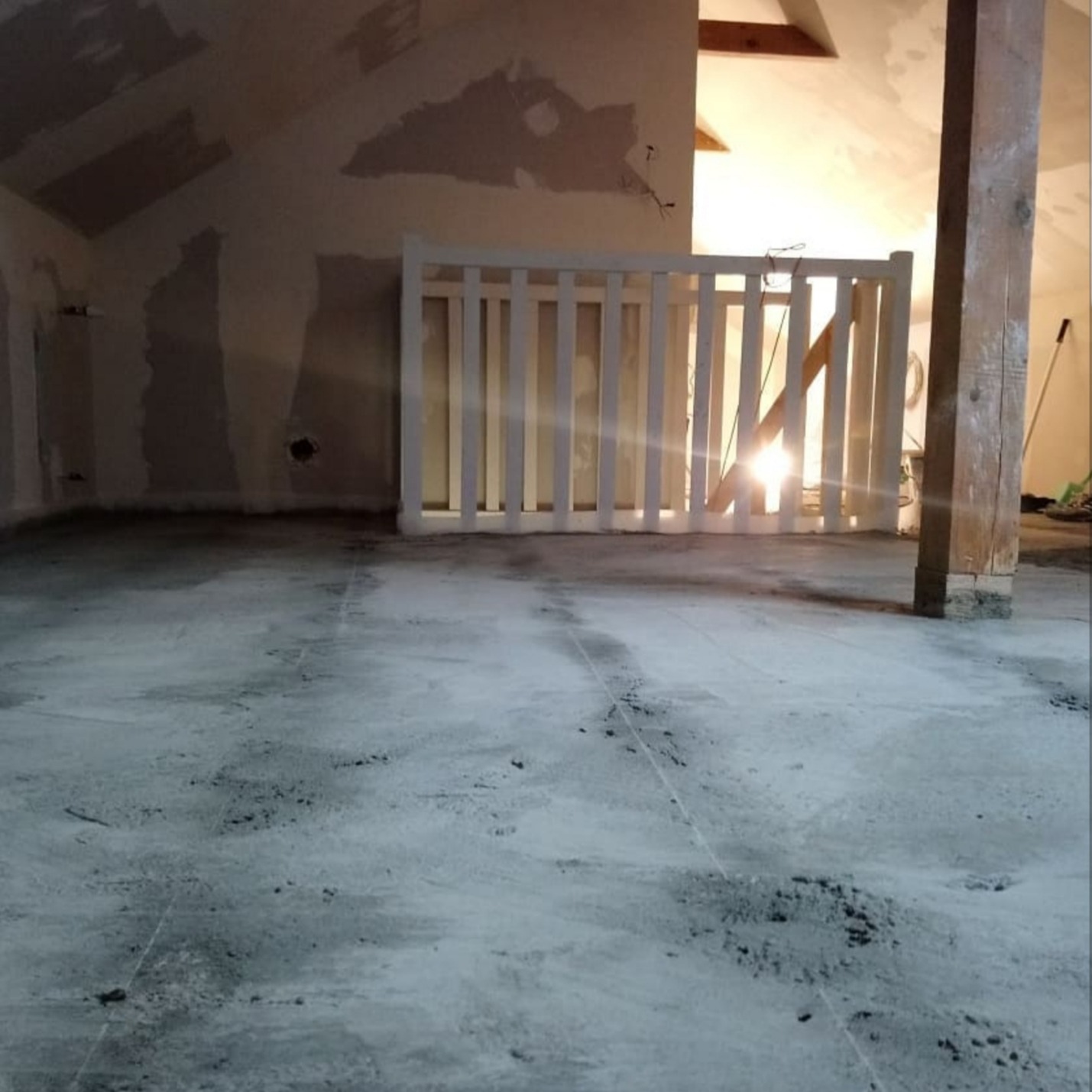 Poddasze w trakcie remontu: wylewka na podłodze, ściany przygotowane do malowania, biała drewniana balustrada, widoczny słup konstrukcyjny.