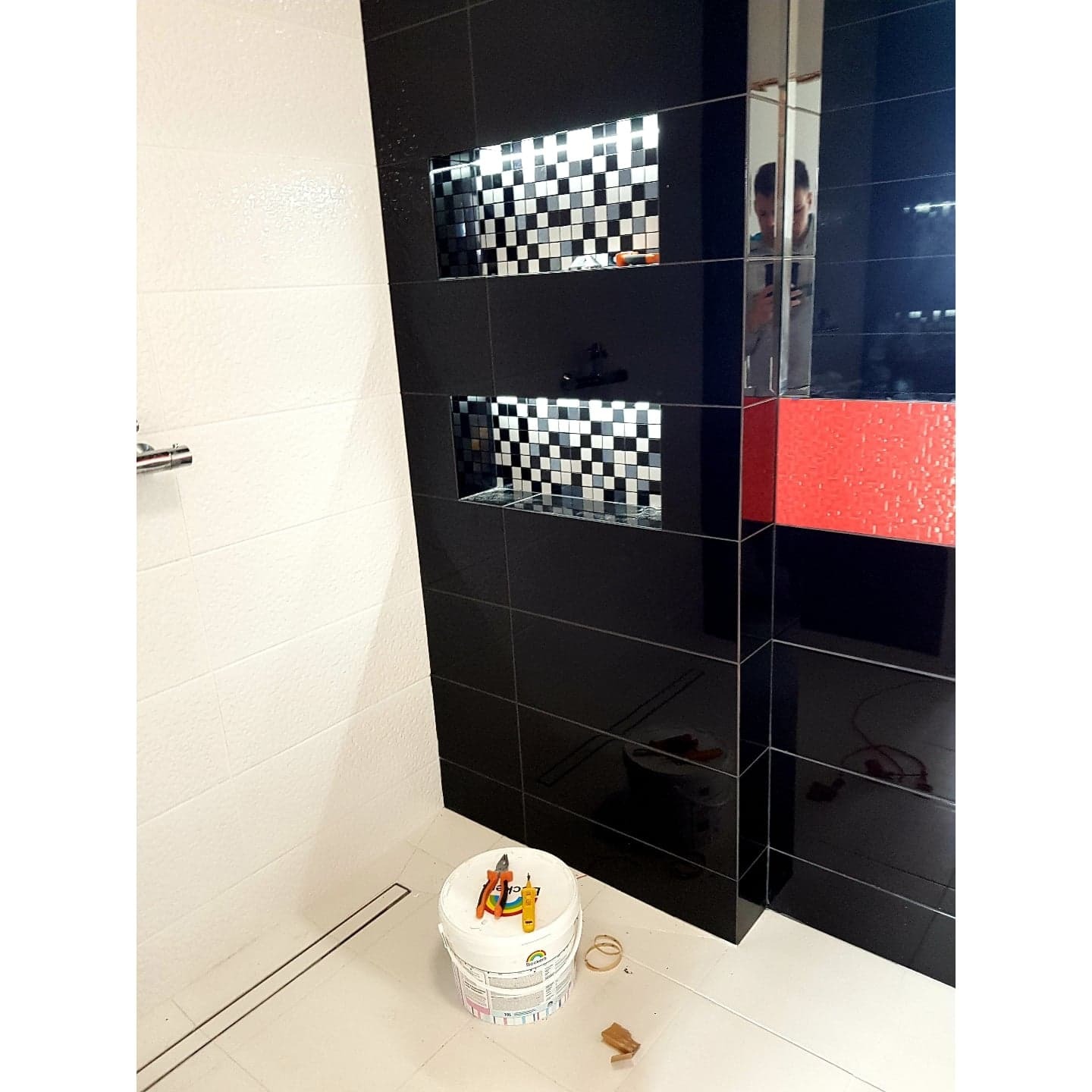 Wykończona łazienka z czarno-białymi płytkami wnękowymi, białe płytki na ścianie i podłodze, wiadro z narzędziami glazurniczymi, widoczne odbicie osoby w lustrzanej płytce.