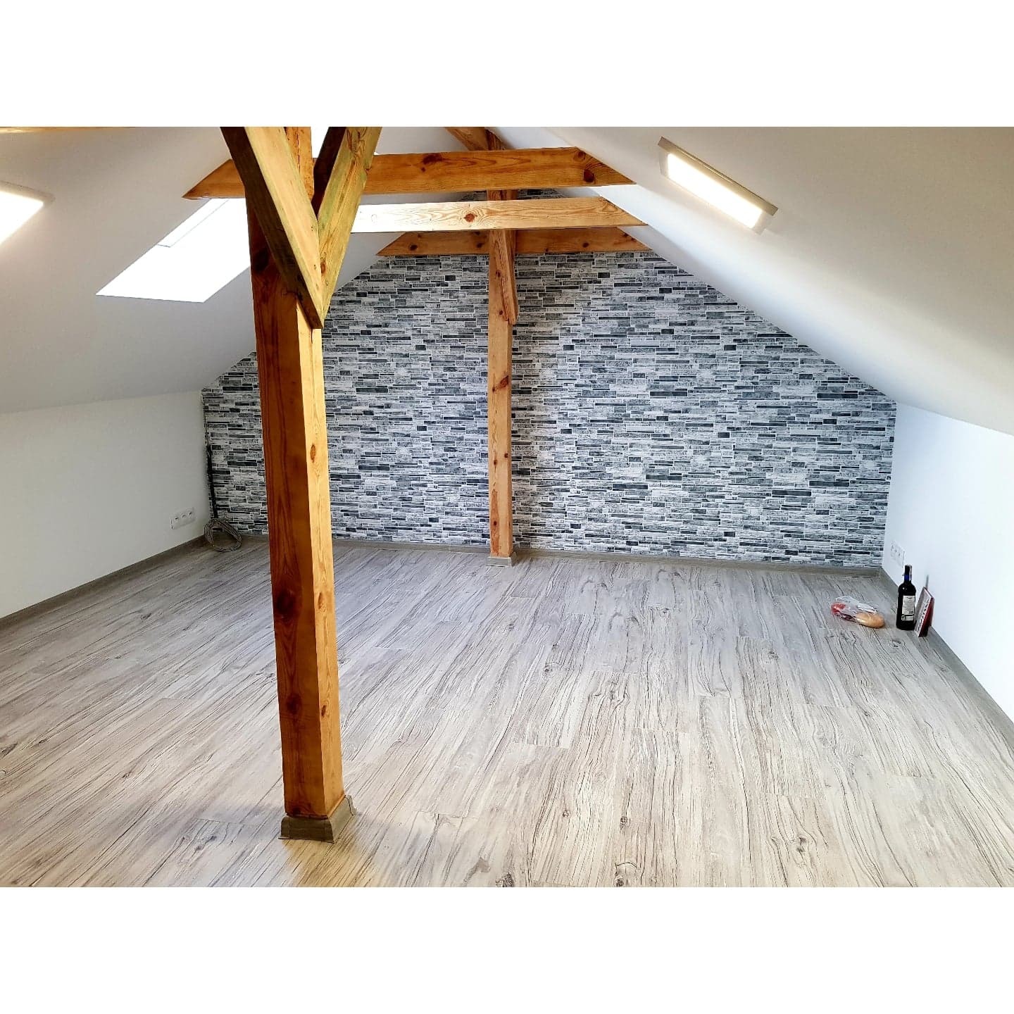 Poddasze po remoncie w Ostrowcu Świętokrzyskim z drewnianymi belkami, wykończone panelami podłogowymi i dekoracyjną okładziną ścienną imitującą kamień, butelka wina i bagietka na podłodze.