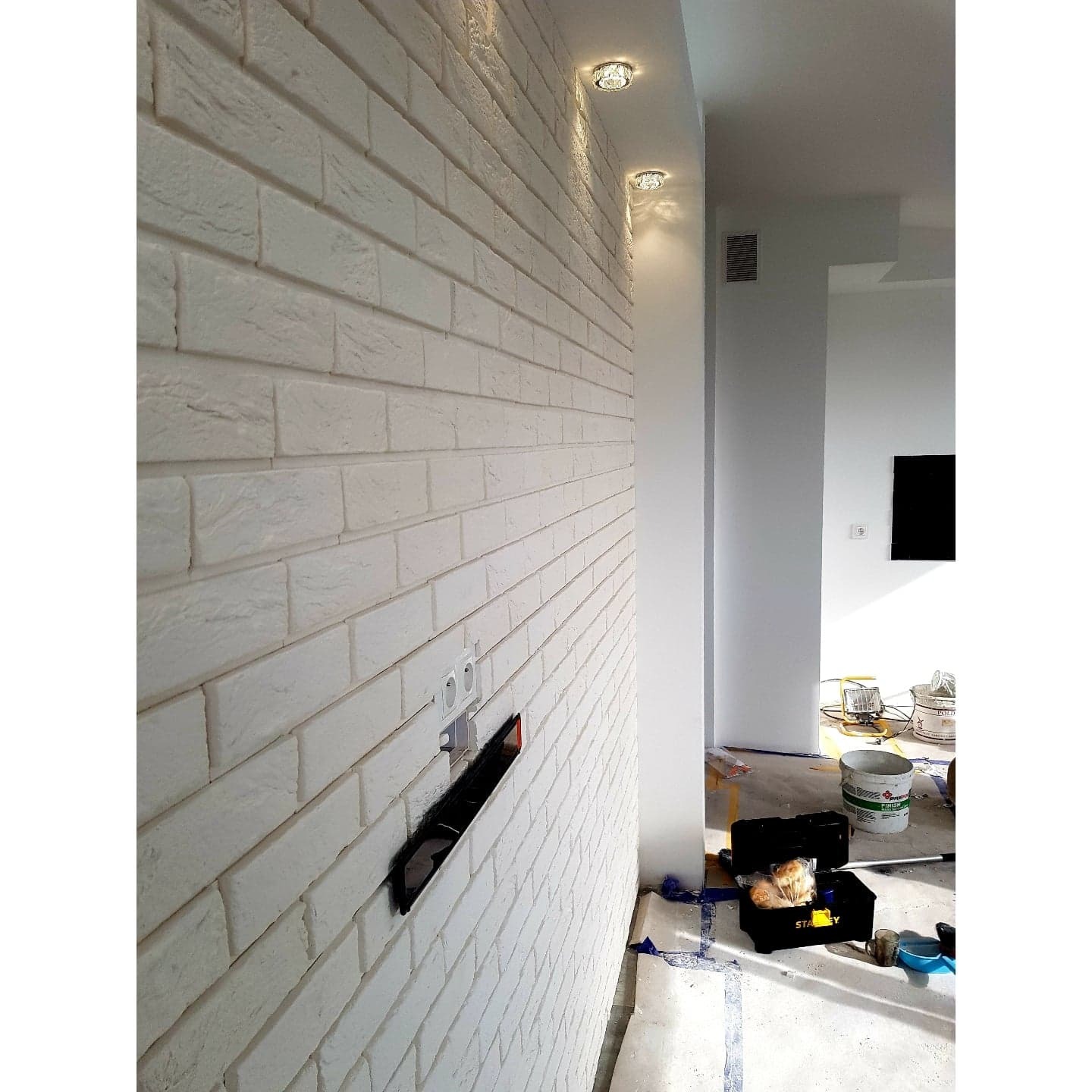 Ściana wykończona białą cegłą dekoracyjną z widocznym otworem na instalację elektryczną, w tle narzędzia i wiadro z farbą, oświetlenie punktowe sufitowe.