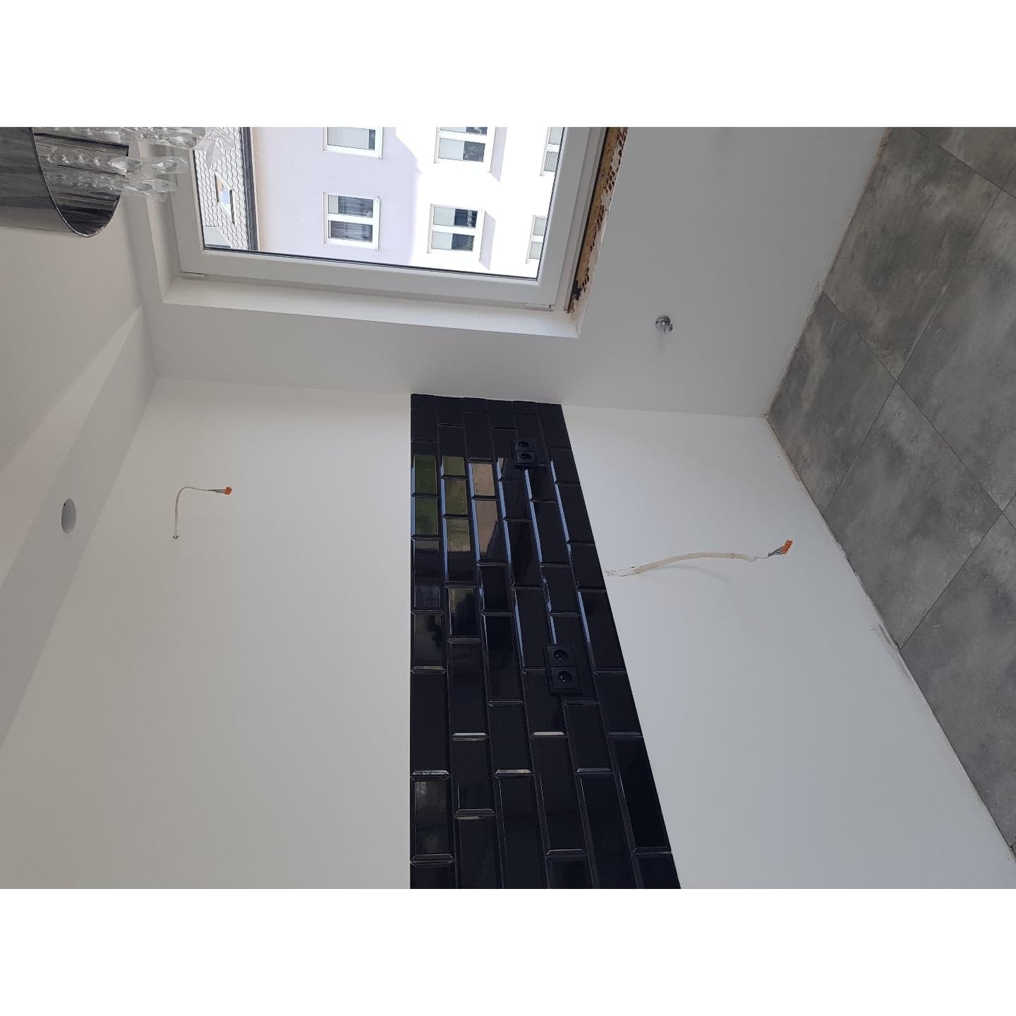 Wnętrze w trakcie prac wykończeniowych: pionowy pas czarnej, błyszczącej glazury na białej ścianie, widoczne gniazdka elektryczne, wystające przewody, szare płytki podłogowe, okno dachowe...