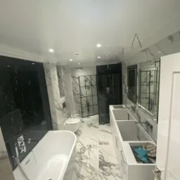 Nowoczesna łazienka z marmurowymi płytkami, czarną kabiną prysznicową typu walk-in i białą wanną wolnostojącą, podwójna umywalka z szafkami, widoczne przewody elektryczne wystające ze ściany.