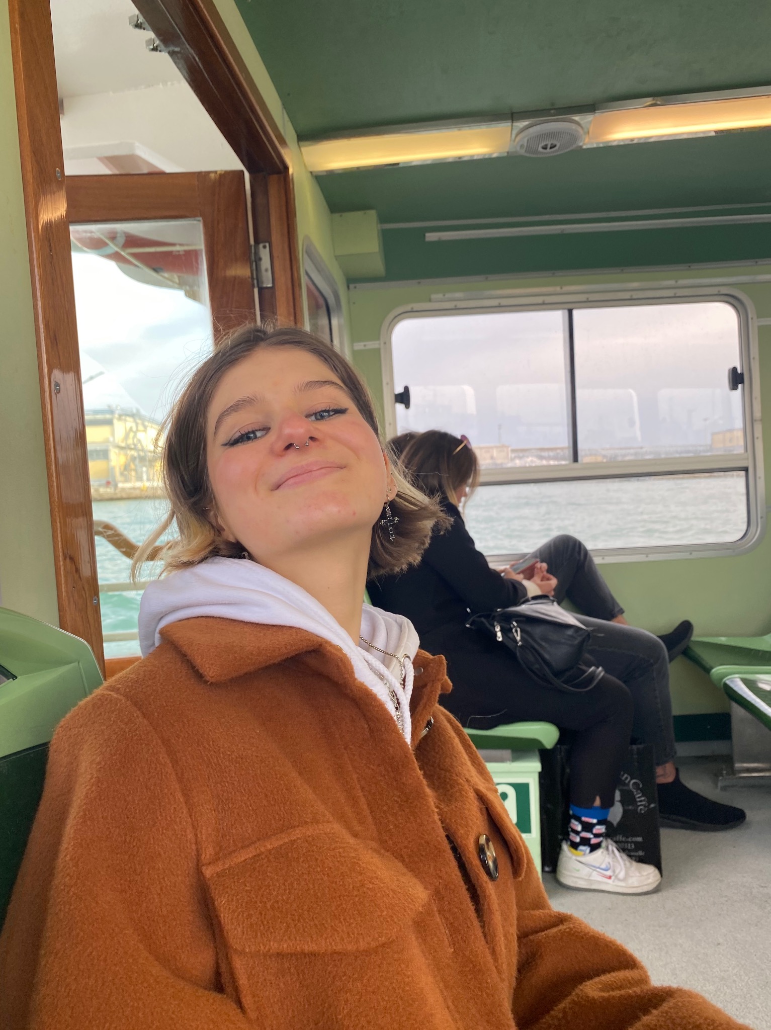 Uśmiechnięta młoda kobieta w brązowej kurtce siedzi wewnątrz promu lub łodzi, w tle widać drugą osobę i zabudowania portowe za oknem.