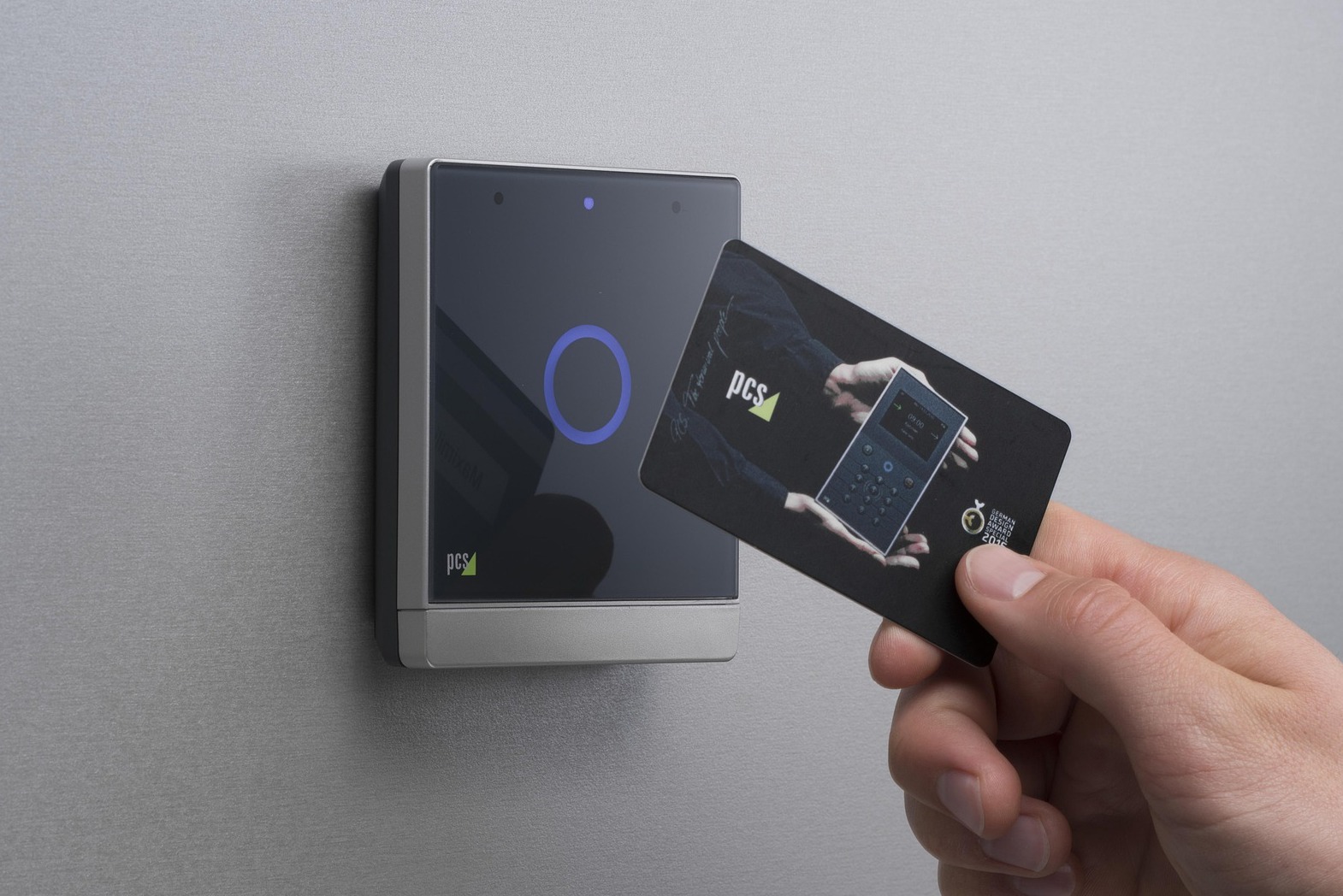 Zbliżenie na czytnik kart zbliżeniowych PCS zamontowany na ścianie, z dłonią przykładającą kartę z nadrukowanym zdjęciem innego czytnika PCS.