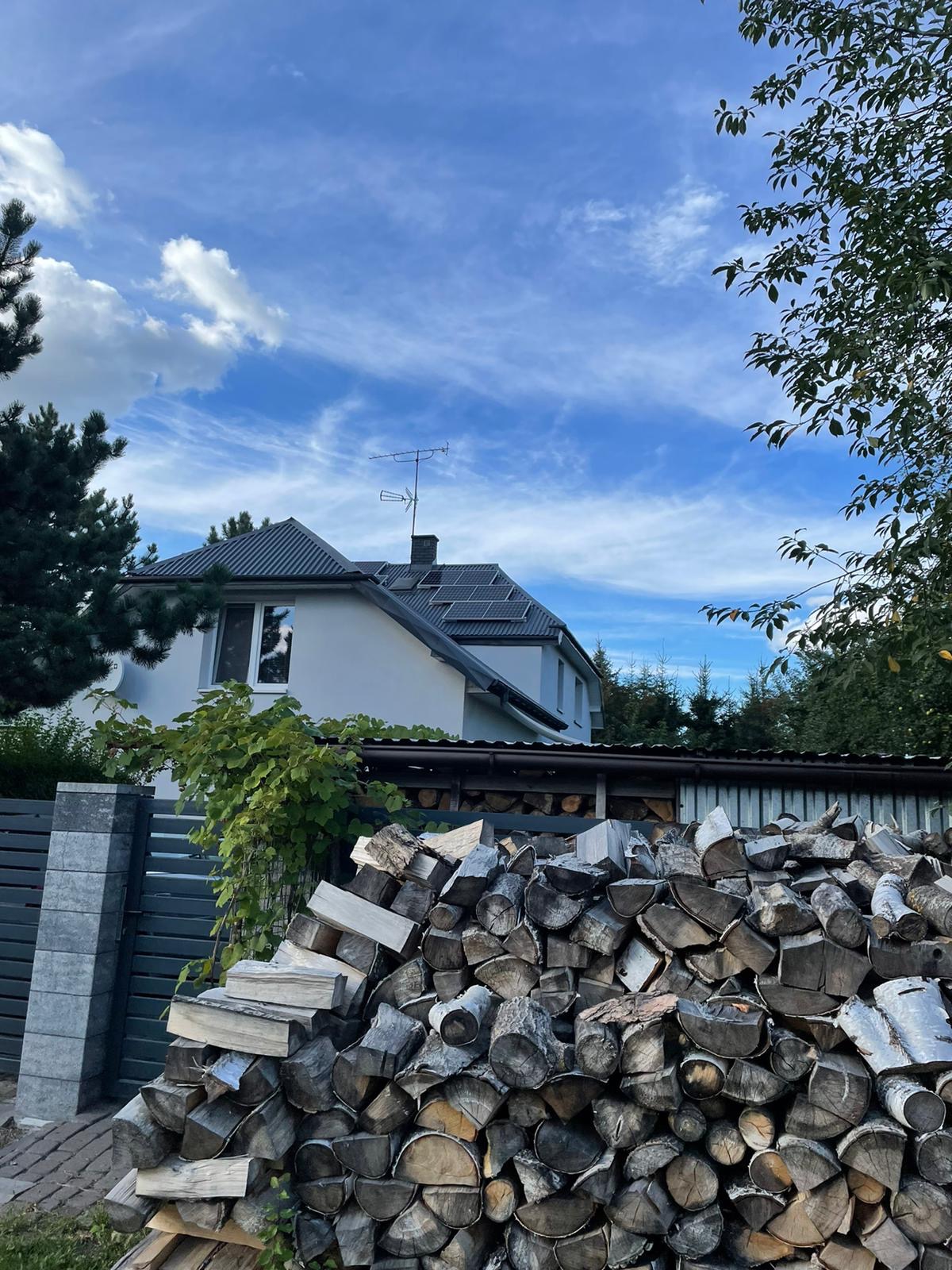 Biały dom jednorodzinny z szarym dachem i panelami słonecznymi, widoczny zza stosu drewna opałowego i szarego ogrodzenia w słoneczny dzień.