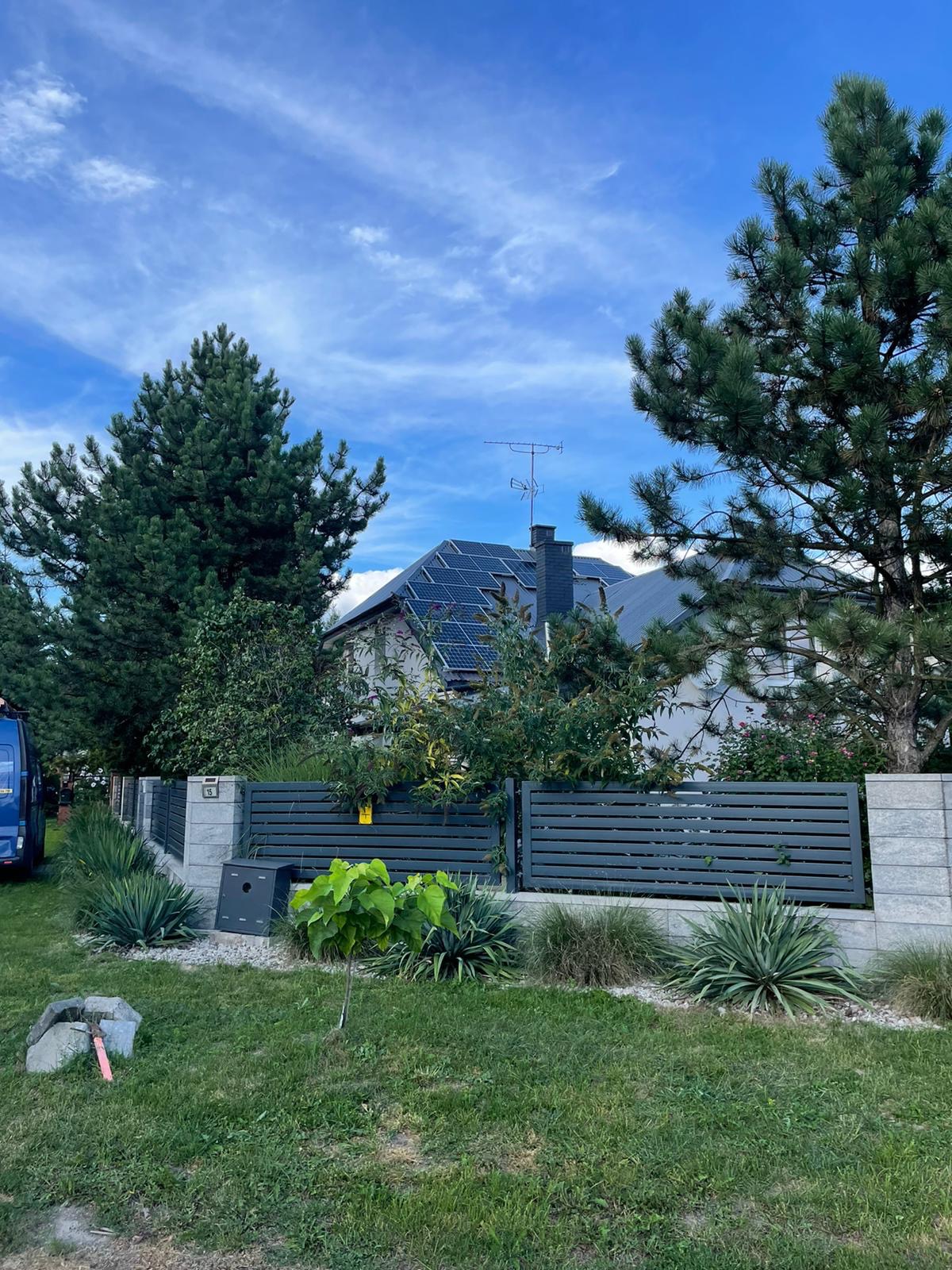 Dom jednorodzinny z szarym dachem pokrytym panelami słonecznymi, widoczny zza szarego ogrodzenia z betonowymi słupkami i bujnej zieleni ogrodu.