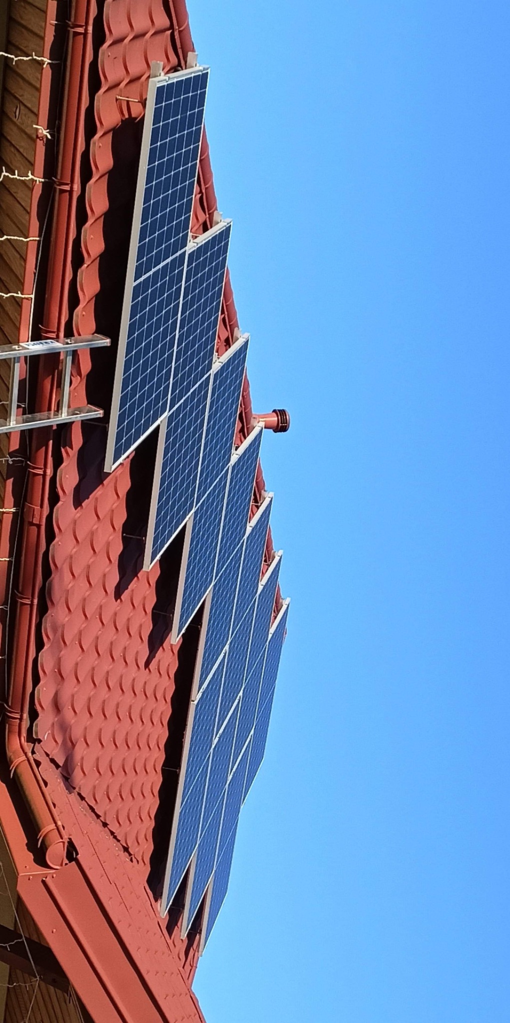 Panele słoneczne zamontowane na czerwonym dachu z dachówki ceramicznej, drabina oparta o ścianę budynku, błękitne niebo w tle.