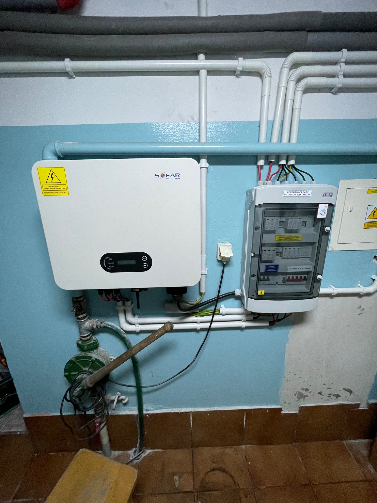 Rozdzielnica elektryczna i inwerter fotowoltaiczny marki Sofar, zamontowane na niebieskiej ścianie w piwnicy, widoczne rury instalacyjne i fragment instalacji wodnej.