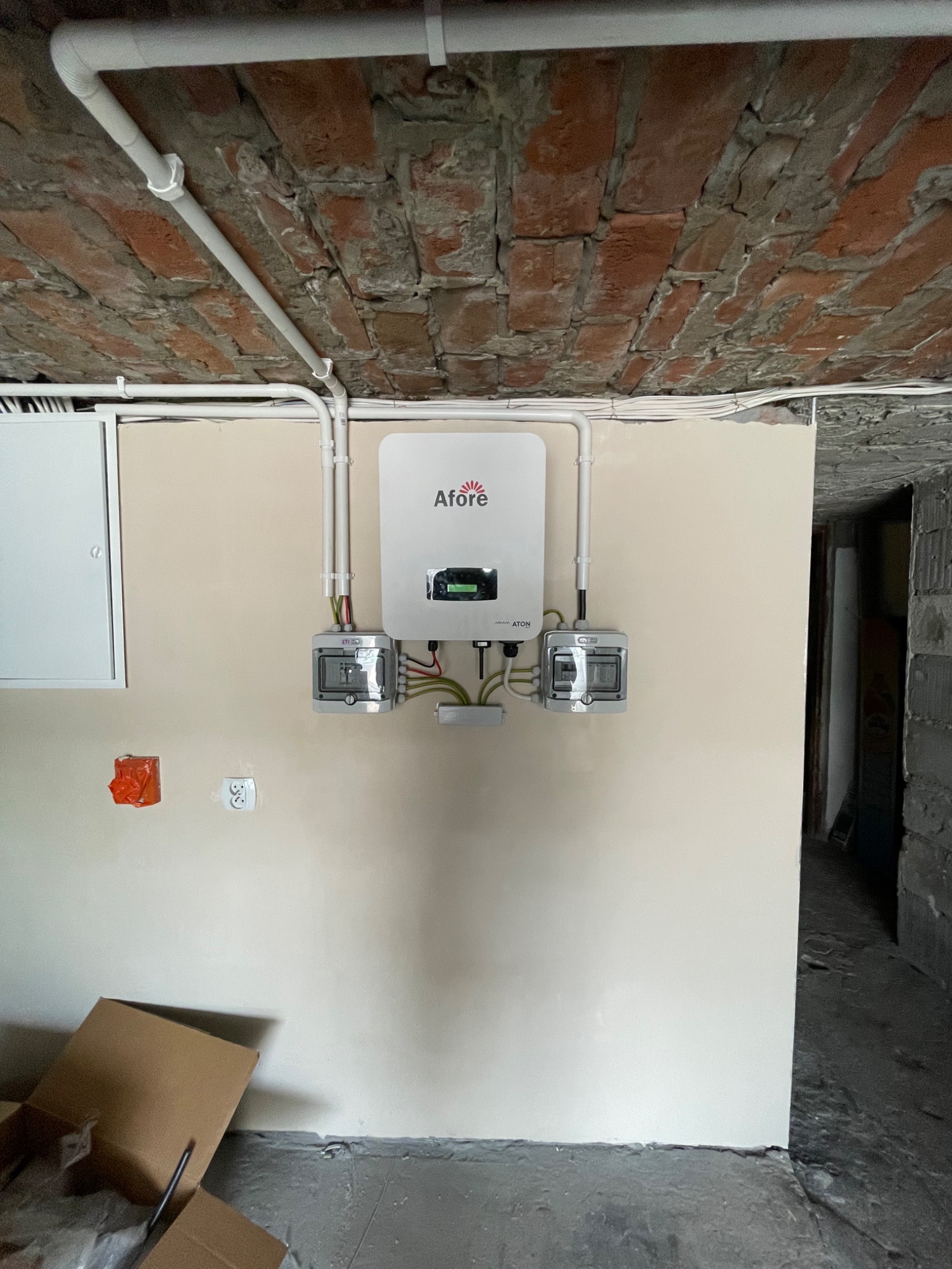 Biały falownik Afore zamontowany na ścianie z widocznymi przewodami, puszkami elektrycznymi i częściowo odsłoniętą cegłą na suficie, w tle fragment otwartego kartonu.