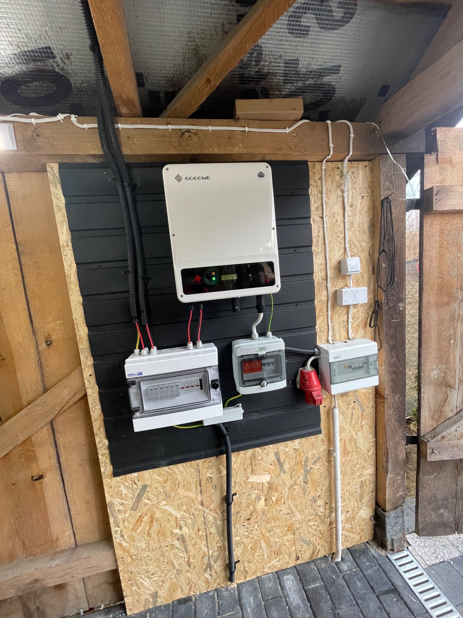Instalacja fotowoltaiczna w drewnianej konstrukcji z widocznym inwerterem GoodWe, rozdzielnicami elektrycznymi i przewodami w peszlach.