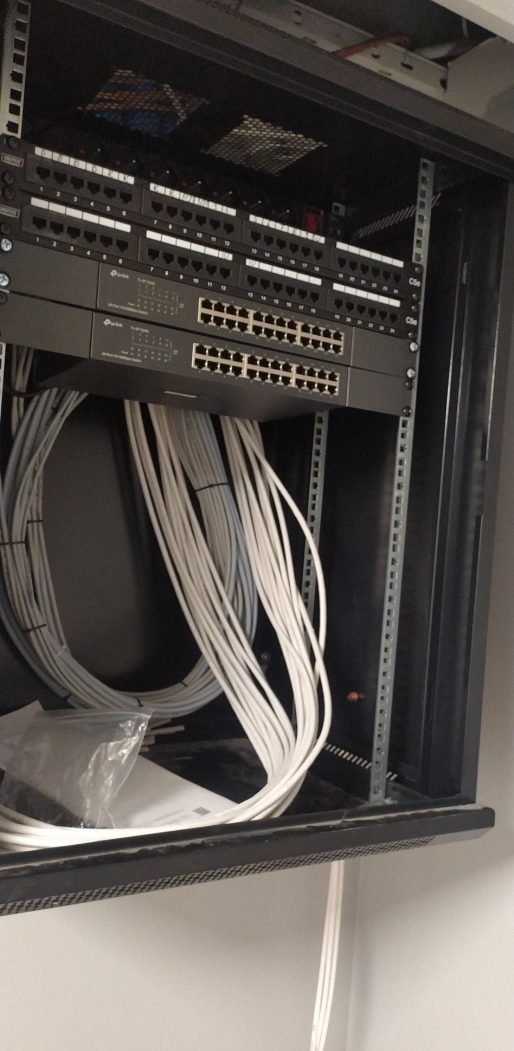 Widok z bliska na szafę rack z patch panelem i switchem, z uporządkowanymi kablami sieciowymi w kolorze białym, w tle widoczna ściana.