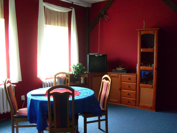 Pokój z czerwonymi ścianami, okrągłym stołem przykrytym niebieskim obrusem, czterema krzesłami, telewizorem i drewnianymi meblami.