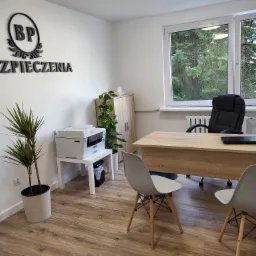 Jasne biuro z biurkiem, krzesłami, roślinami doniczkowymi i napisem 'Ubezpieczenia' na ścianie, widok z okna na zieleń.