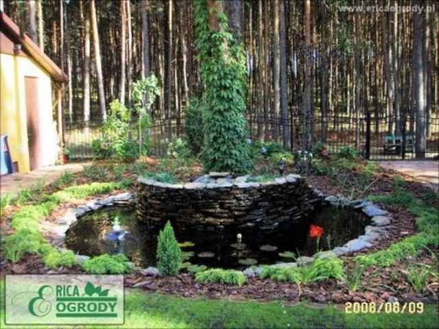 Oczko wodne z kamienną kaskadą i roślinnością ozdobną, otoczone leśnym krajobrazem, z widocznym logo firmy ogrodniczej w rogu zdjęcia.