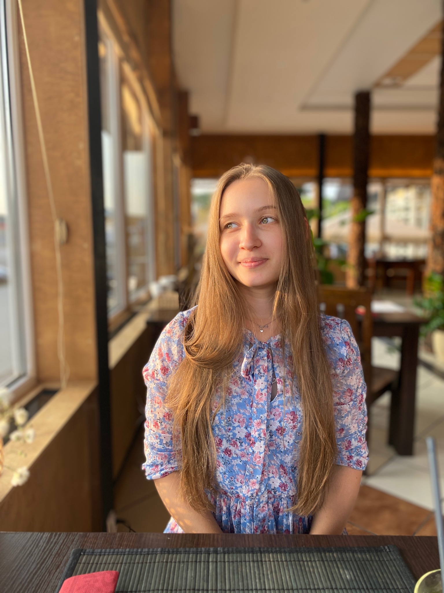 Portret młodej kobiety w kwiecistej sukience, siedzącej przy stole w kawiarni, z widokiem na zewnątrz przez okno.